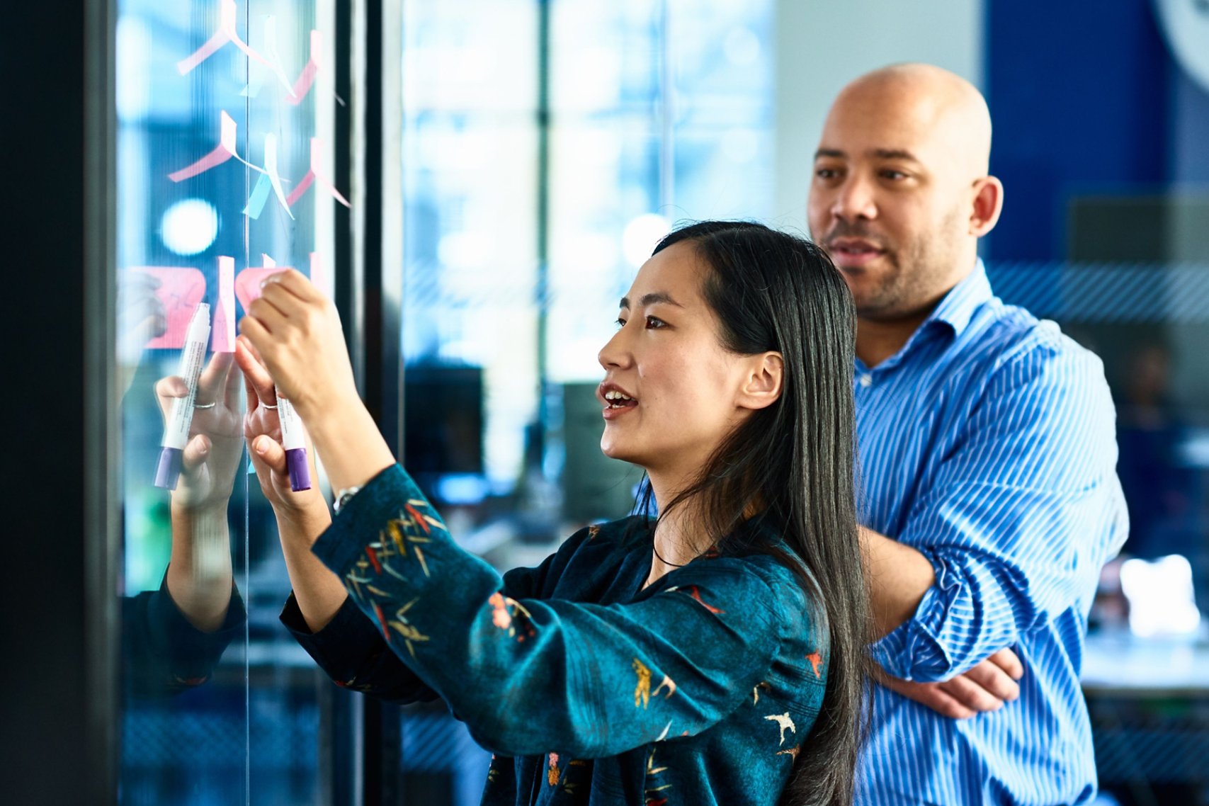 A woman is collaborating with a man by writing notes on a glass wall in an office