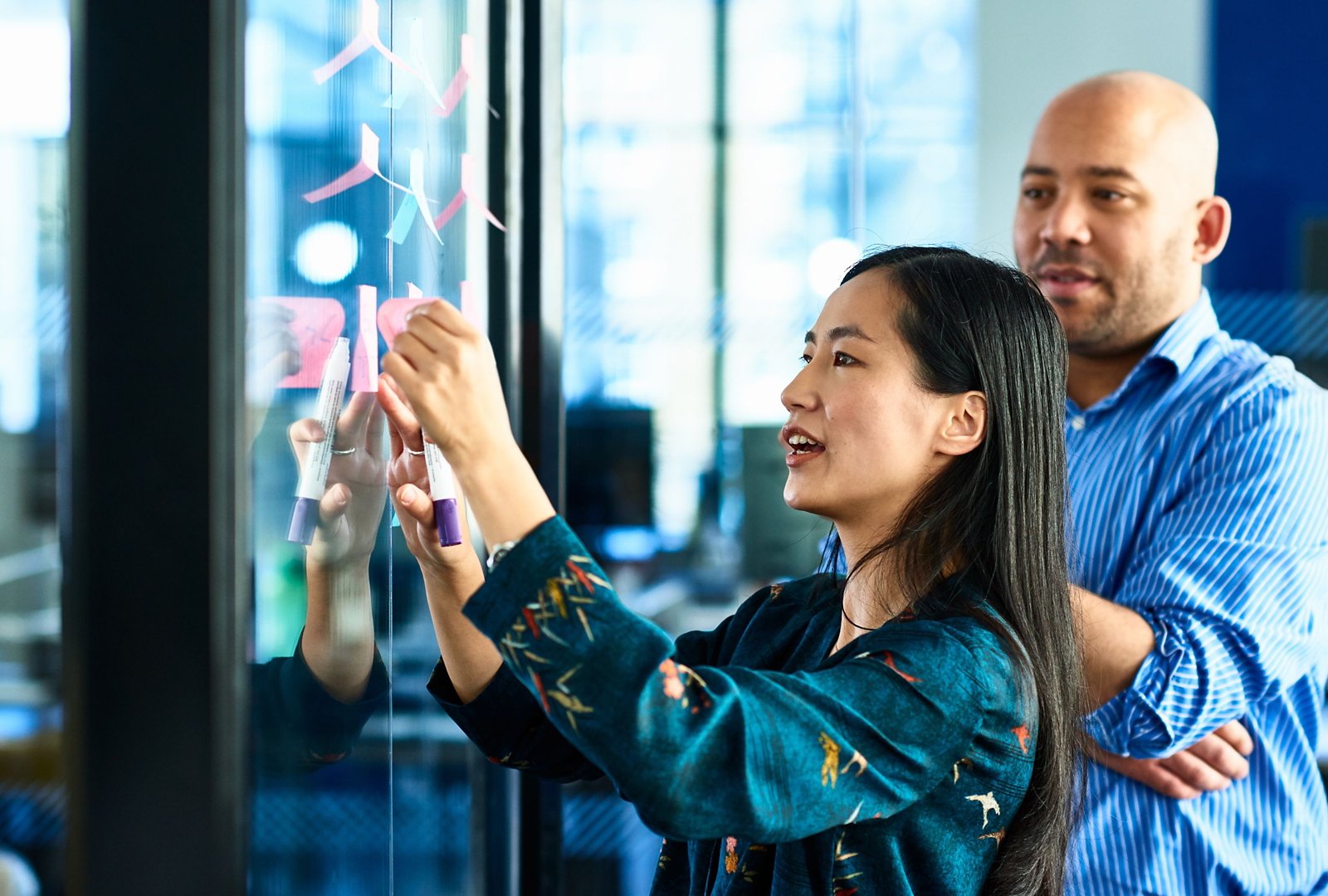 A woman is collaborating with a man by writing notes on a glass wall in an office