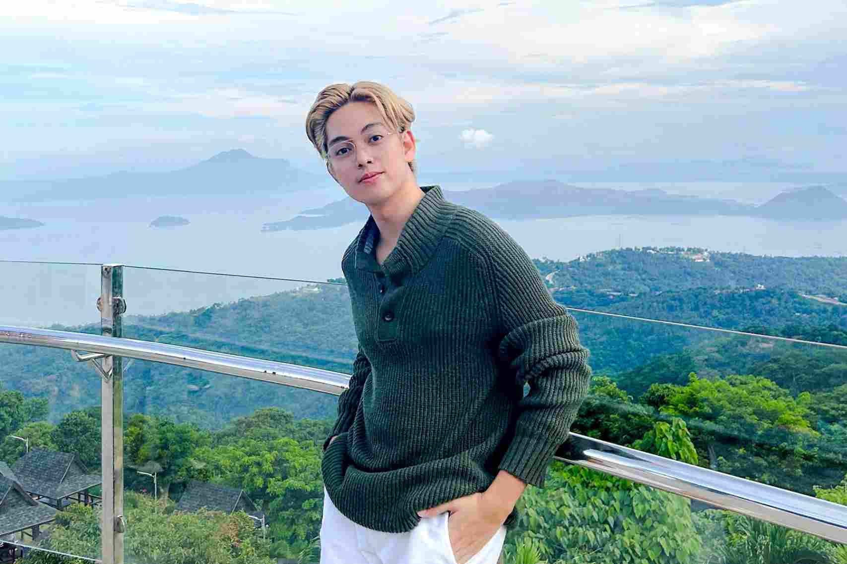 A man with light blond hair wearing green sweater standing at a glass railing