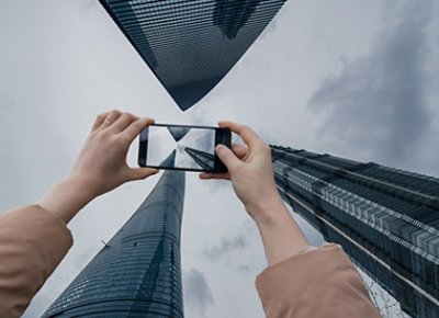 An image shows a hand using cellphone capturing the buildings towards the sky