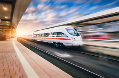White modern speed train in motion on railway station at sunset. Train on railroad track with motion blur effect in Europe in evening. Railway platform. Industrial landscape. Railway tourism