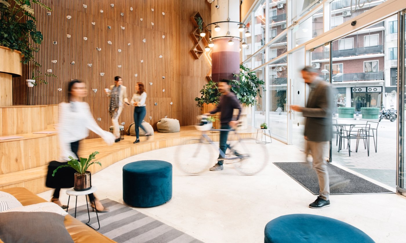 Modern lobby with wooden walls, plants, glass windows, and people walking and cycling.