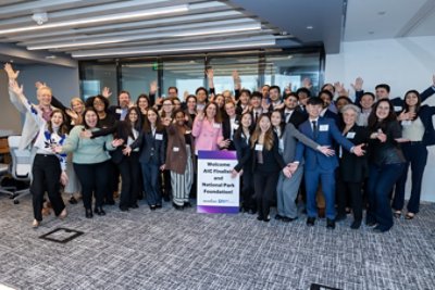 A large group of professionally dressed adults poses together in a modern office setting, smiling and waving enthusiastically. The group includes men and women of various ages and backgrounds, gathered around a sign that reads 'Welcome AIC Finalists and National Park Foundation!' The atmosphere is lively and positive, with bright lighting and contemporary decor. The event appears to be a formal or semi-formal gathering, emphasizing teamwork and achievement.