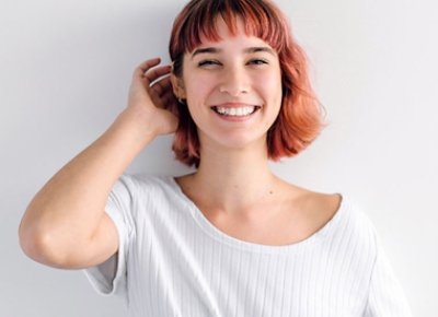 Colored hair woman smiling with white shirt on and leaning on a while wall