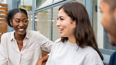 Two women smiling at each other outside the building with a man figure in the blur background