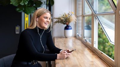 Woman with earphone and holding the phone sitting near the window looking outside smiling