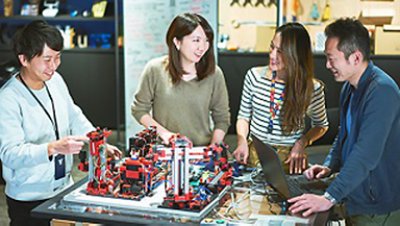 Two men and two women working at a table and smiling, chips and wires and connected devides configuration on the table