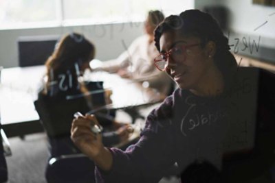 A woman writing on a mirrored board with her teammate at the background