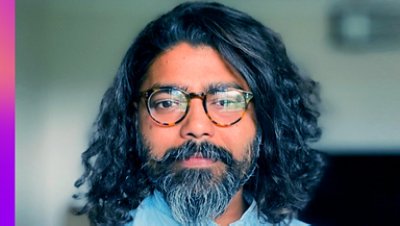 A face of a bearded long-haired man with eyeglasses