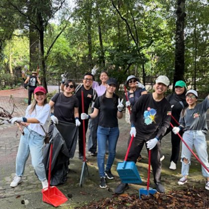 A group of volunteers takes part in an outdoor clean-up activity in a green, wooded setting. Participants are pictured together wearing protective gloves and holding cleaning equipment, including brooms, dustpans and waste bags. The image captures colleag