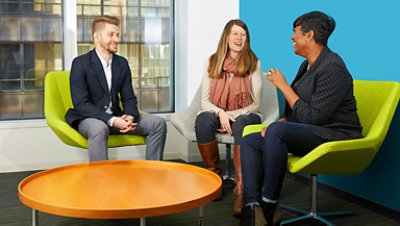 Three colleagues sitting and discussing while smiling, in front of them is a round orange table.