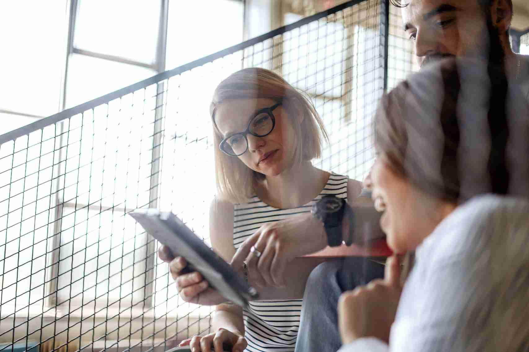 A woman wearing glasses and a striped top holds a digital tablet, leading a discussion on work or data with two colleagues on an industrial-style staircase