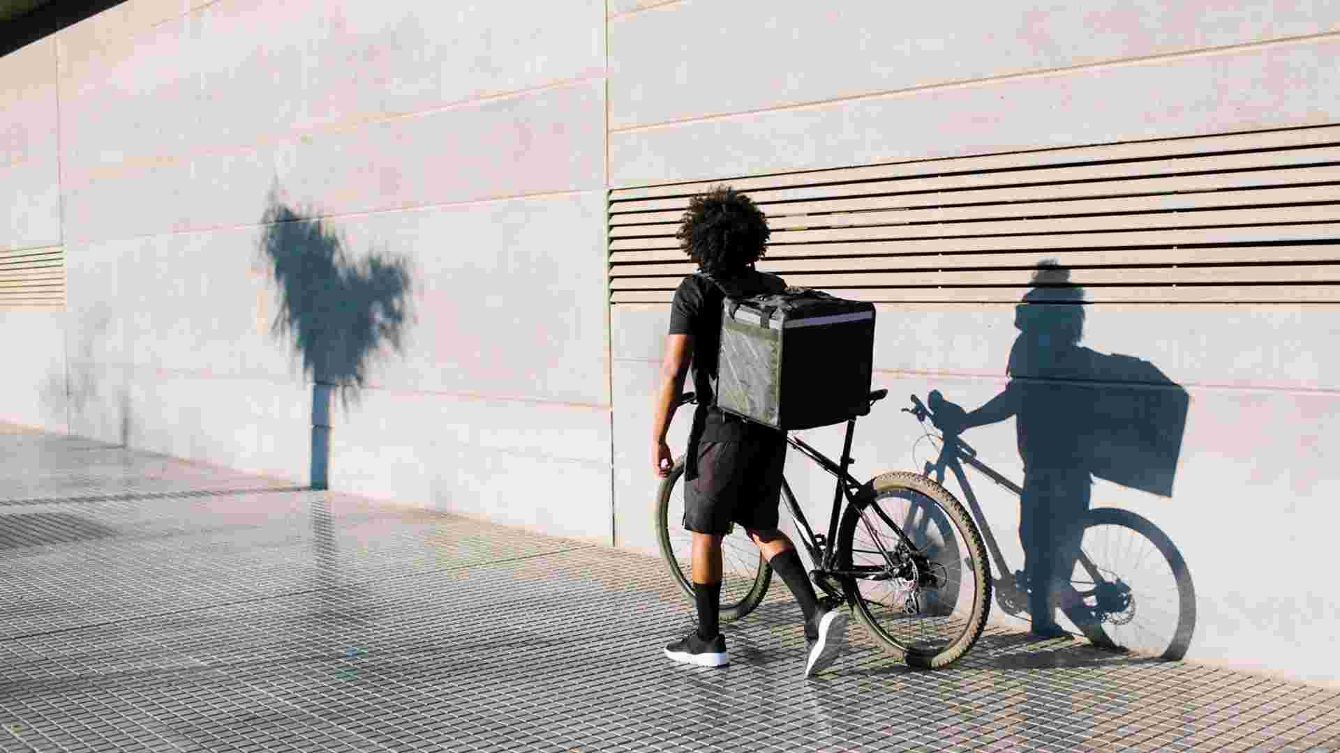 Back view of black delivery rider with box and bike walking on city pavement against wall with shadow while looking away in sunlight