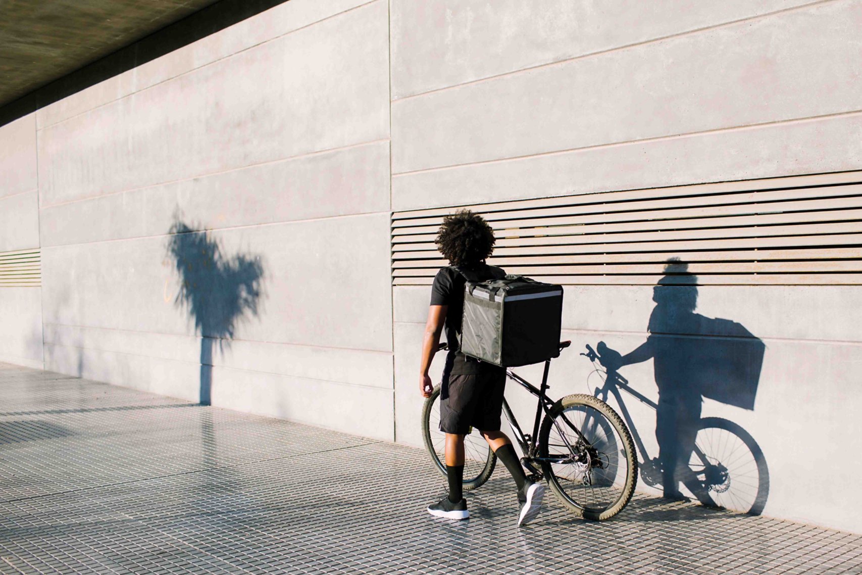 Back view of delivery rider with box and bike walking on city pavement against wall with shadow while looking away in sunlight