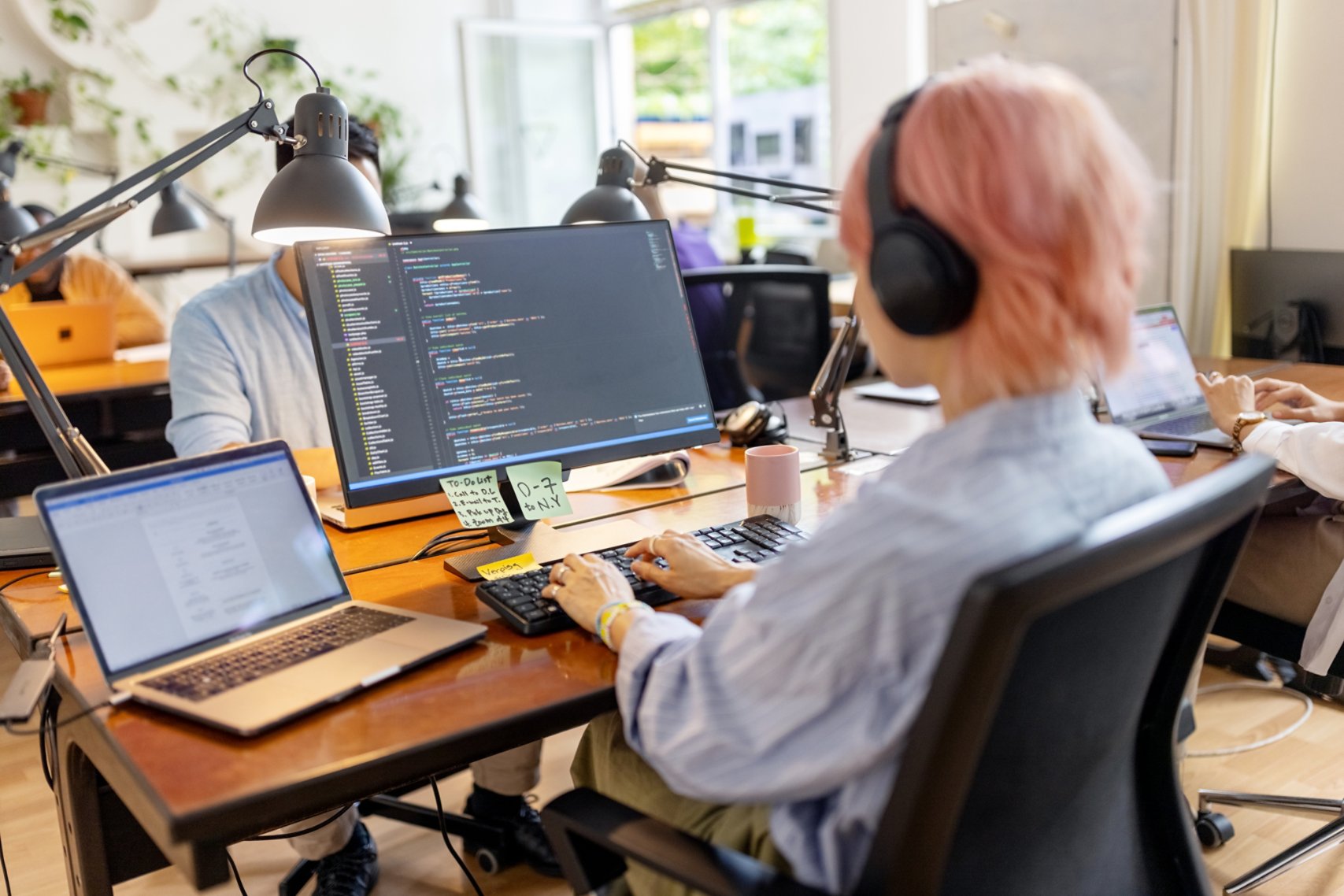 Rear view of a female computer programmer working at her desk at a startup company. Businesswoman wearing a headset coding on computer at coworking office space.