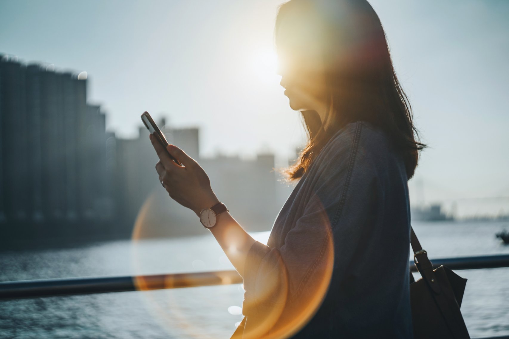 Silhouette of woman using smartphone by commercial dock against sunlight with lens flare in city